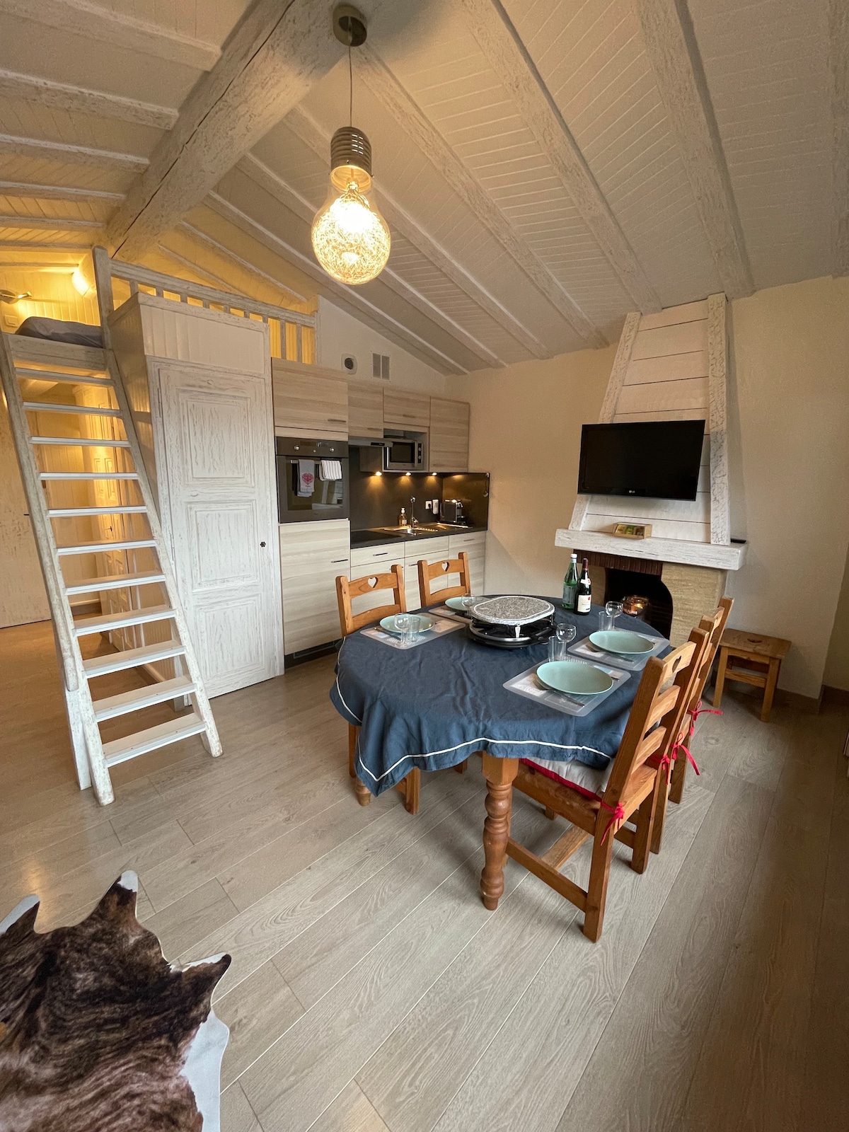 The open living area features a wooden dining table set for four, with a dark grey tablecloth. A kitchen area is equipped with modern appliances, including a stovetop and microwave. A staircase leads to the lofted sleeping area. A cozy seating area includes a television and a decorative fireplace.