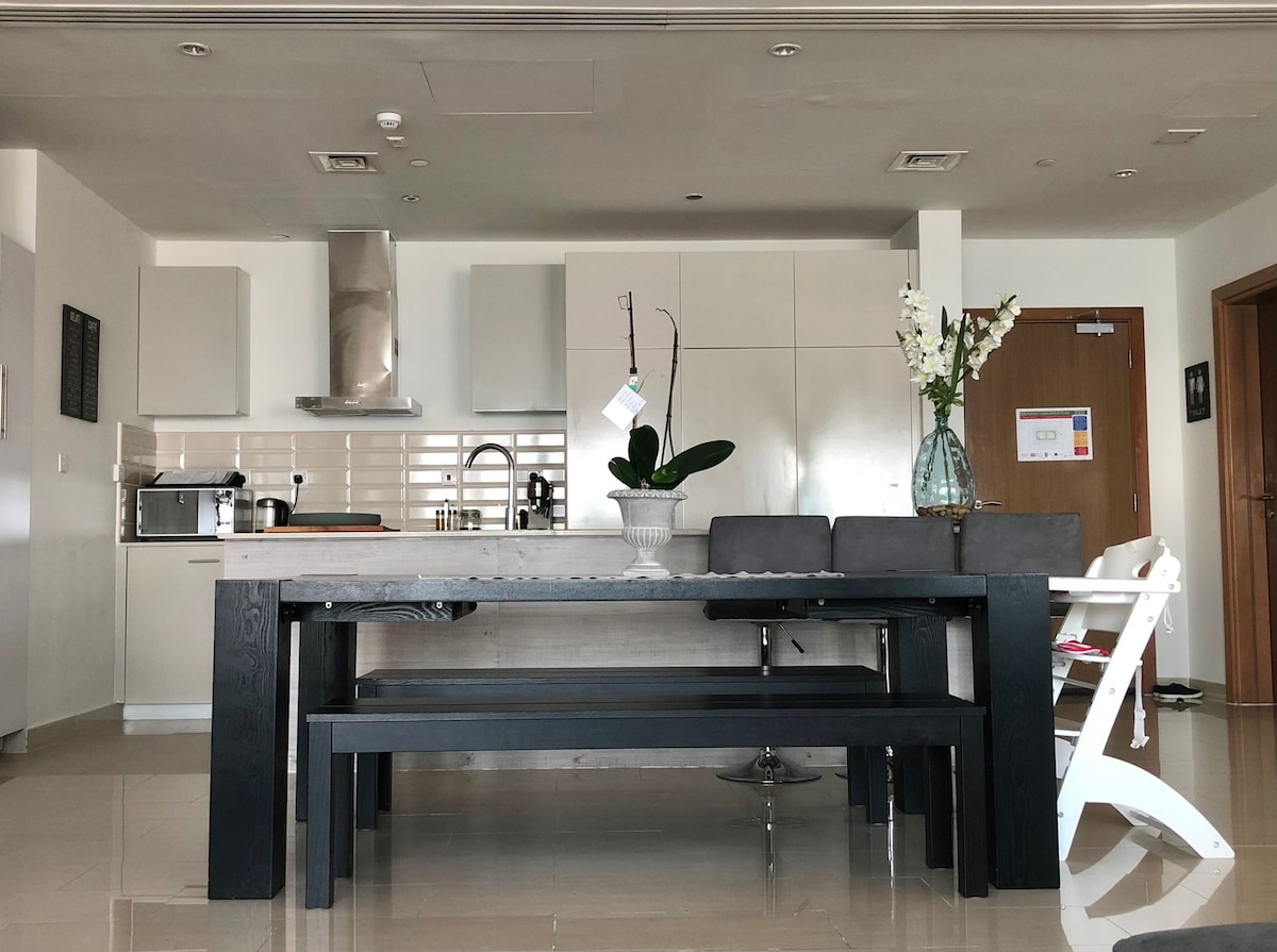 A modern kitchen area features a large wooden dining table accompanied by sleek black benches. Subtle decorative elements, including a vase with flowers, enhance the space, while stainless steel appliances and white cabinetry create a clean and functional environment.