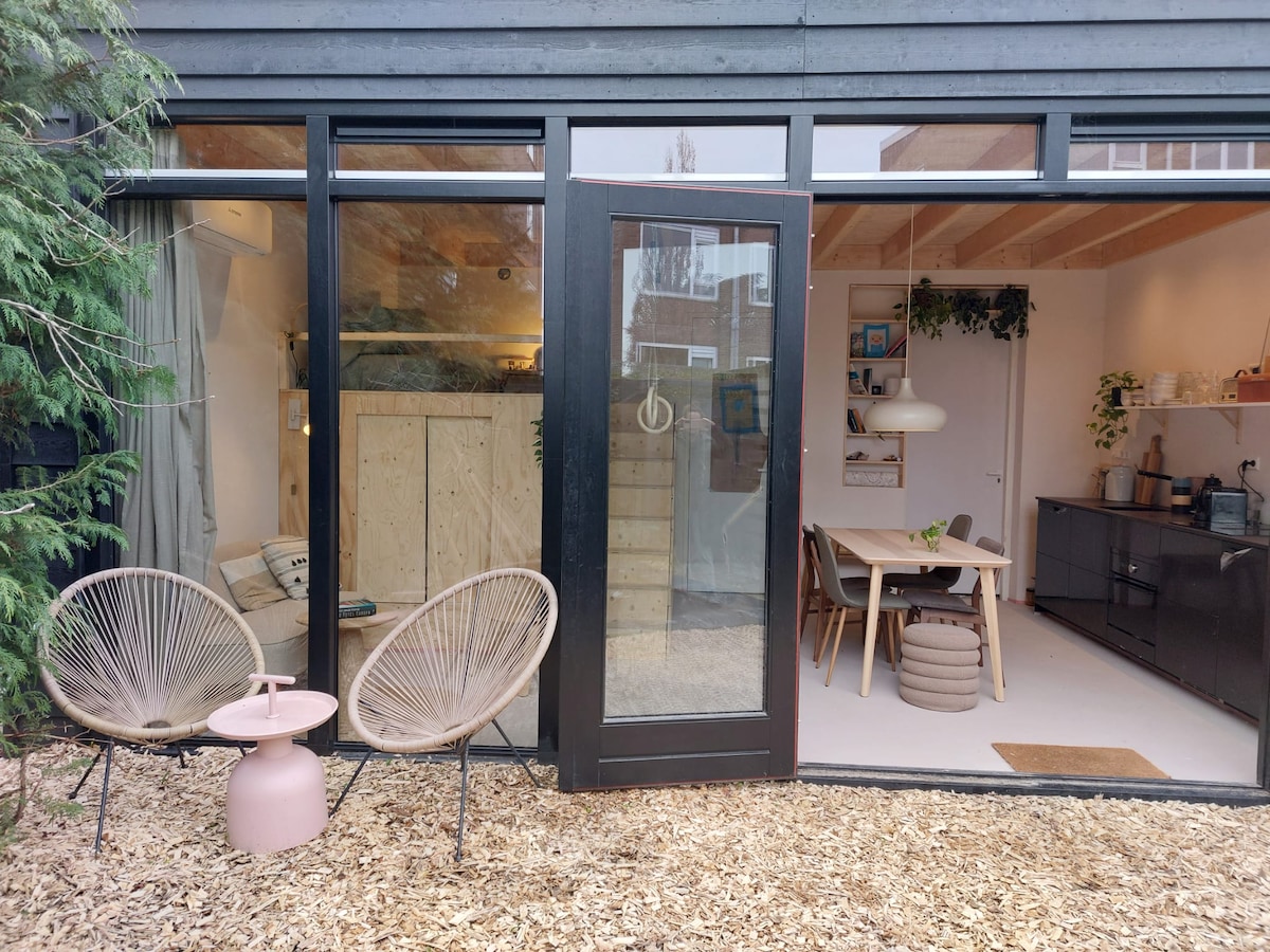 The entrance to a modern studio is framed by glass doors. Two stylish chairs are positioned outside, accompanied by a small table. The interior reveals a dining table and a well-equipped kitchen, with ample natural light illuminating the space.