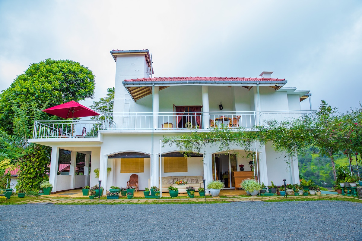 The two-story building presents a clean white facade complemented by a red roof. A spacious balcony is visible on the upper level, adorned with seating and an umbrella. Lush greenery surrounds the property, enhancing the tranquil atmosphere.