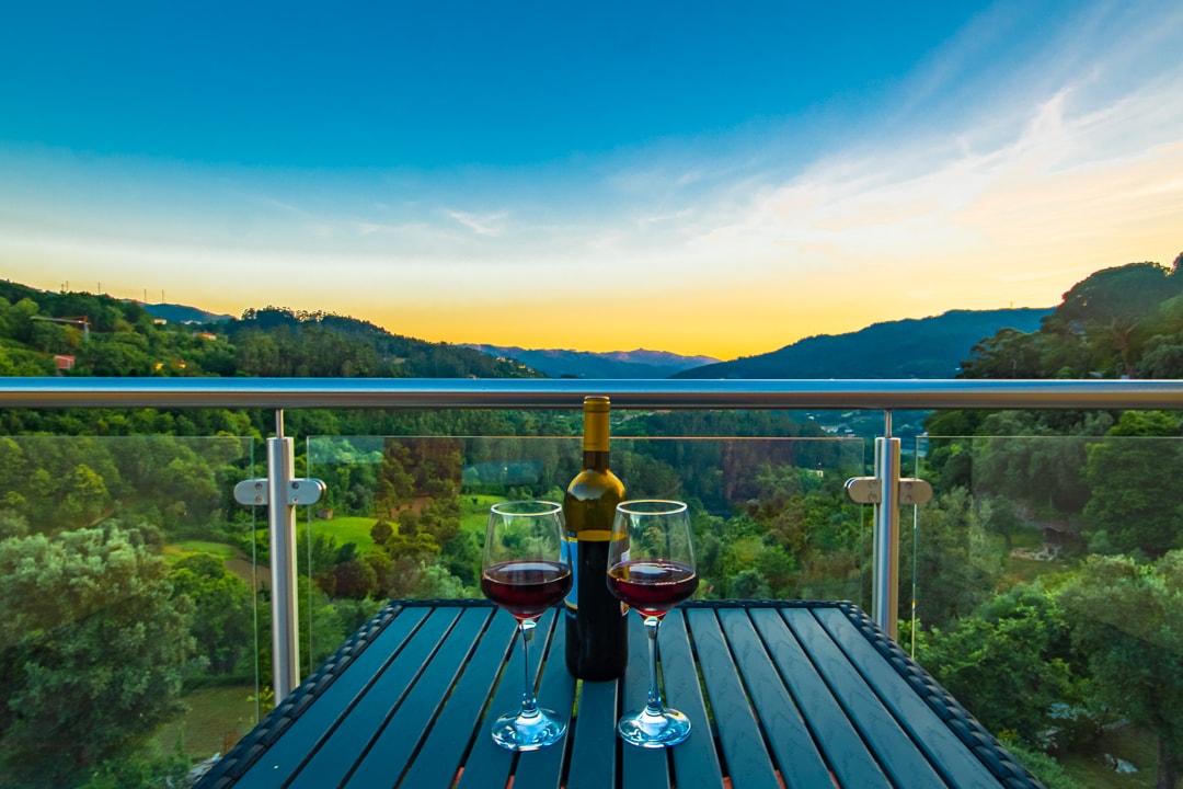 A set table with two glasses filled with red wine and a bottle placed between them is displayed on a balcony. Rolling hills and valleys are visible in the background, under a clear sky transitioning to sunset hues.