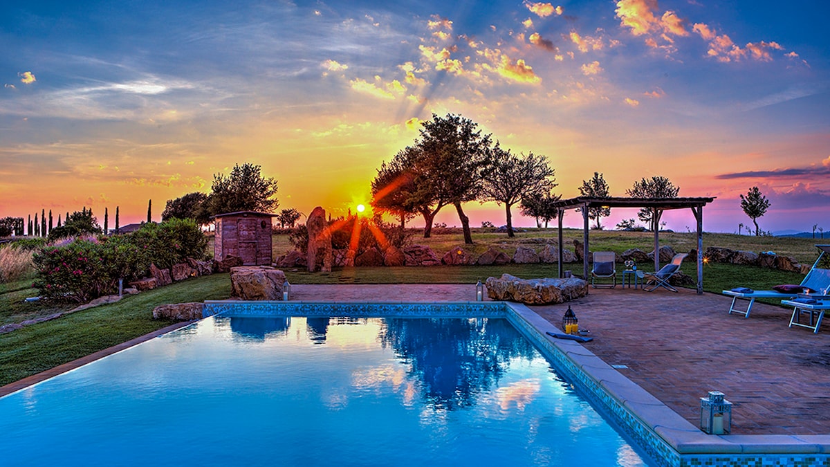 A tranquil outdoor space features a glistening pool reflecting a vibrant sunset sky. Soft silhouettes of trees and rustic structures are visible, surrounded by lush greenery and neatly arranged lounge chairs. The warm hues of dusk create a serene atmosphere.