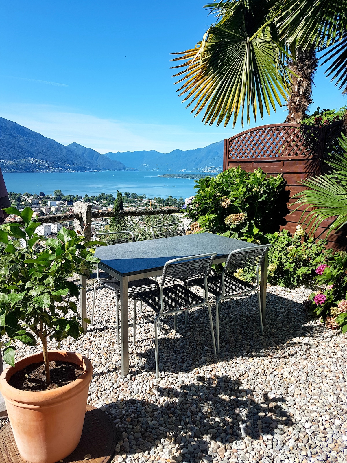 A garden terrace features a rectangular table surrounded by four metal chairs, overlooking a view of Lago Maggiore and the surrounding mountains. Lush greenery and potted plants add a vibrant touch to the outdoor space, creating a serene environment.