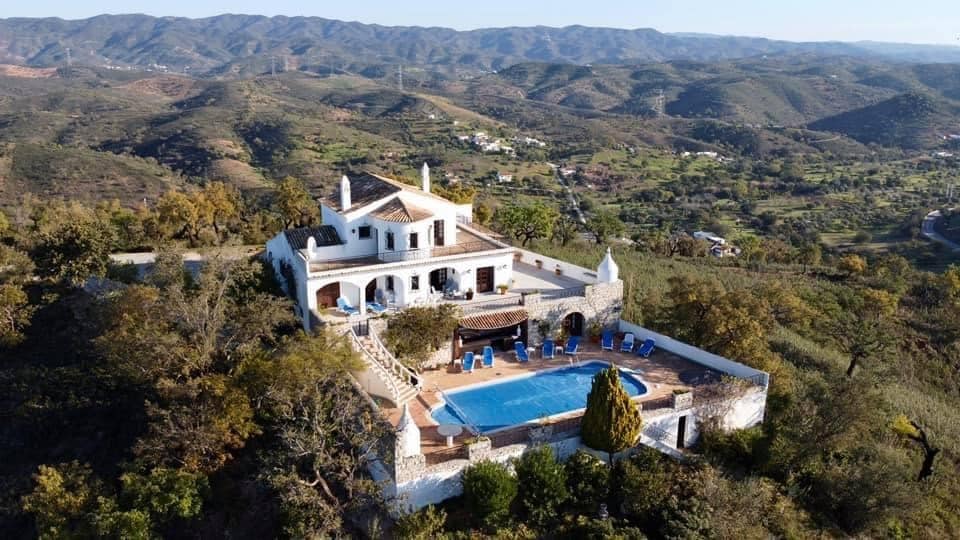 An aerial view of a spacious villa perched on a hillside, surrounded by rolling green hills. A large blue pool is positioned in the foreground, with several sun loungers arranged around it. The multi-level structure features terraced balconies, white walls, and red-tiled roofing.