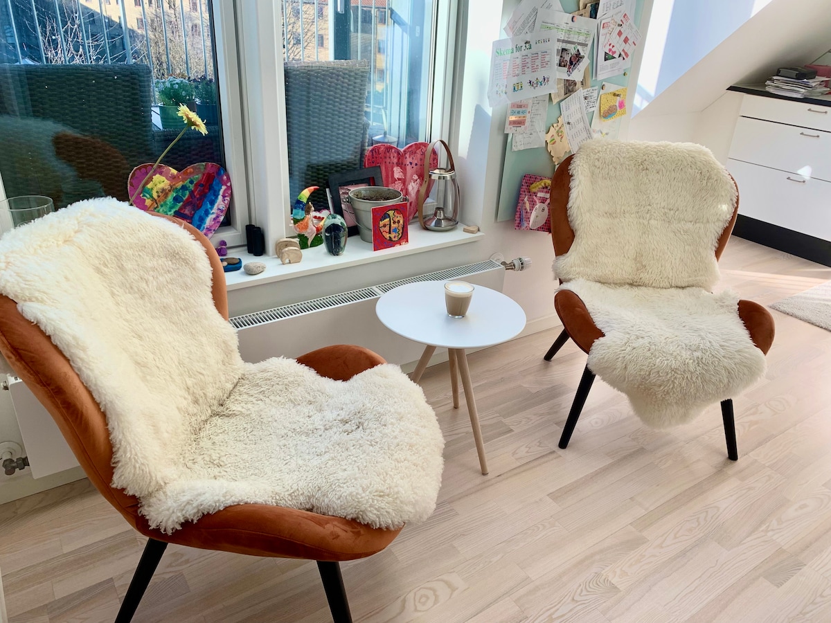 Two cozy chairs with soft, textured coverings are positioned near a window, inviting natural light. A small, round table is situated between them, complementing the earthy tones. Nearby, personal items and colorful artwork are displayed on the wall, adding a touch of character to the space.