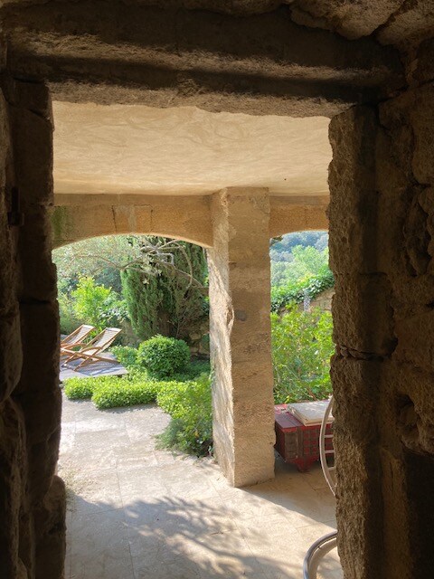 A stone archway frames a view of a serene outdoor space, featuring a well-maintained garden with lush greenery. Two lounge chairs can be seen on a patio area, offering a peaceful retreat surrounded by nature.