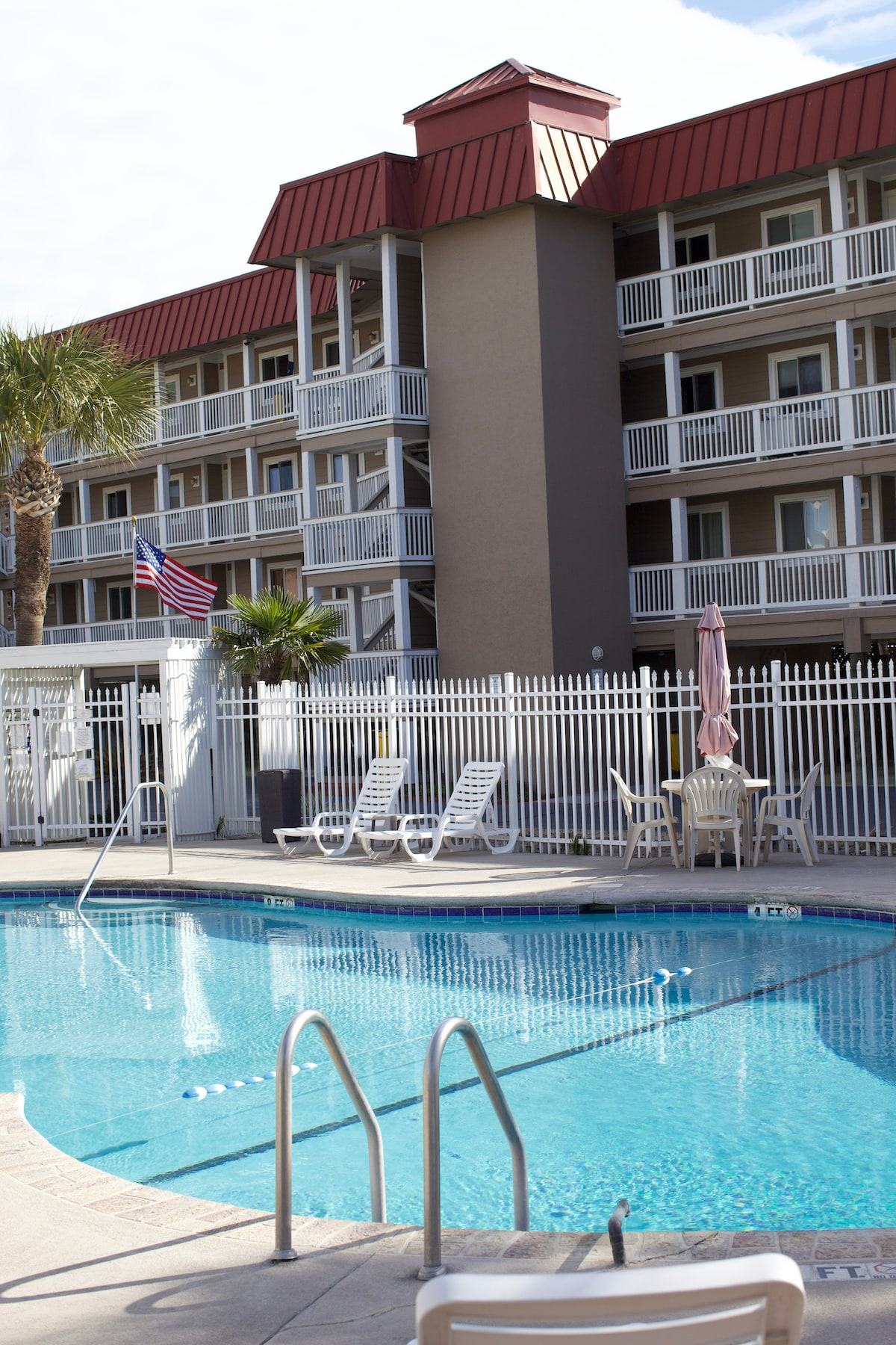 A clean swimming pool is framed by tall palm trees and a white picket fence. Multiple lounge chairs are arranged around the pool area, with the building facade visible in the background. An American flag is displayed prominently near the entrance of the pool.