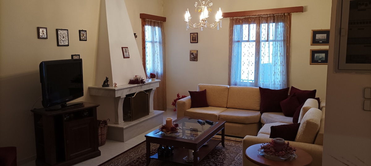 A comfortable living room features a light-colored sofa arranged around a wooden coffee table. A cozy fireplace is located against one wall, complemented by framed pictures and decorative items. Natural light filters through sheer curtains at two windows, enhancing the inviting atmosphere.