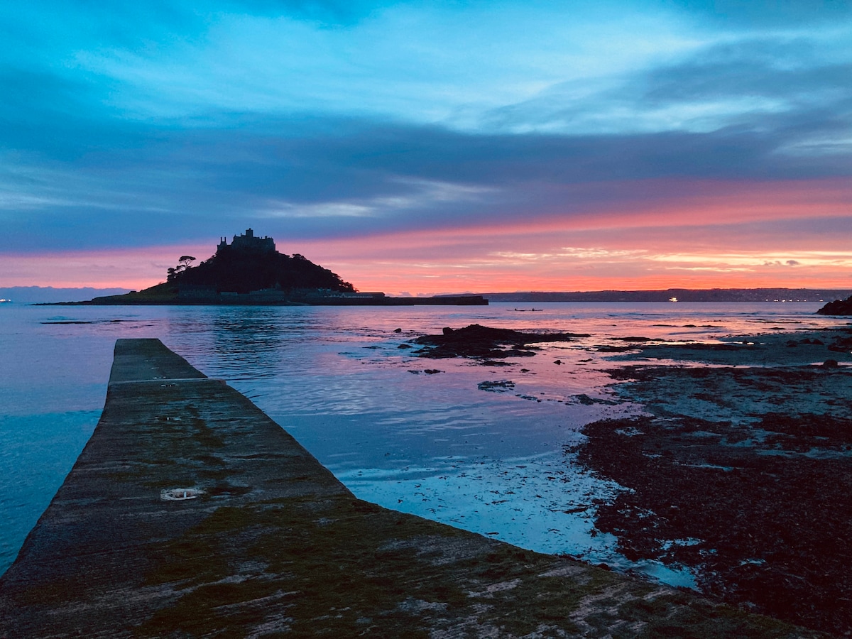 A serene coastal scene captures a long stone pier extending over calm waters. A silhouette of a distant island with a castle structure is visible against a vibrant sunset sky, transitioning from blue to warm hues. Reflections of the colorful sky shimmer on the water's surface.