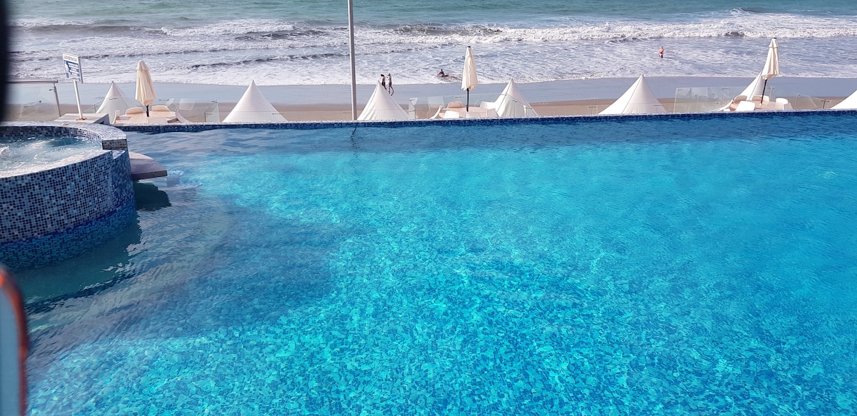 An infinity pool reflects the bright blue sky, blending seamlessly with the ocean waves in the distance. Beach umbrellas and lounge chairs are arranged along the sandy shore, creating a relaxed seaside atmosphere.