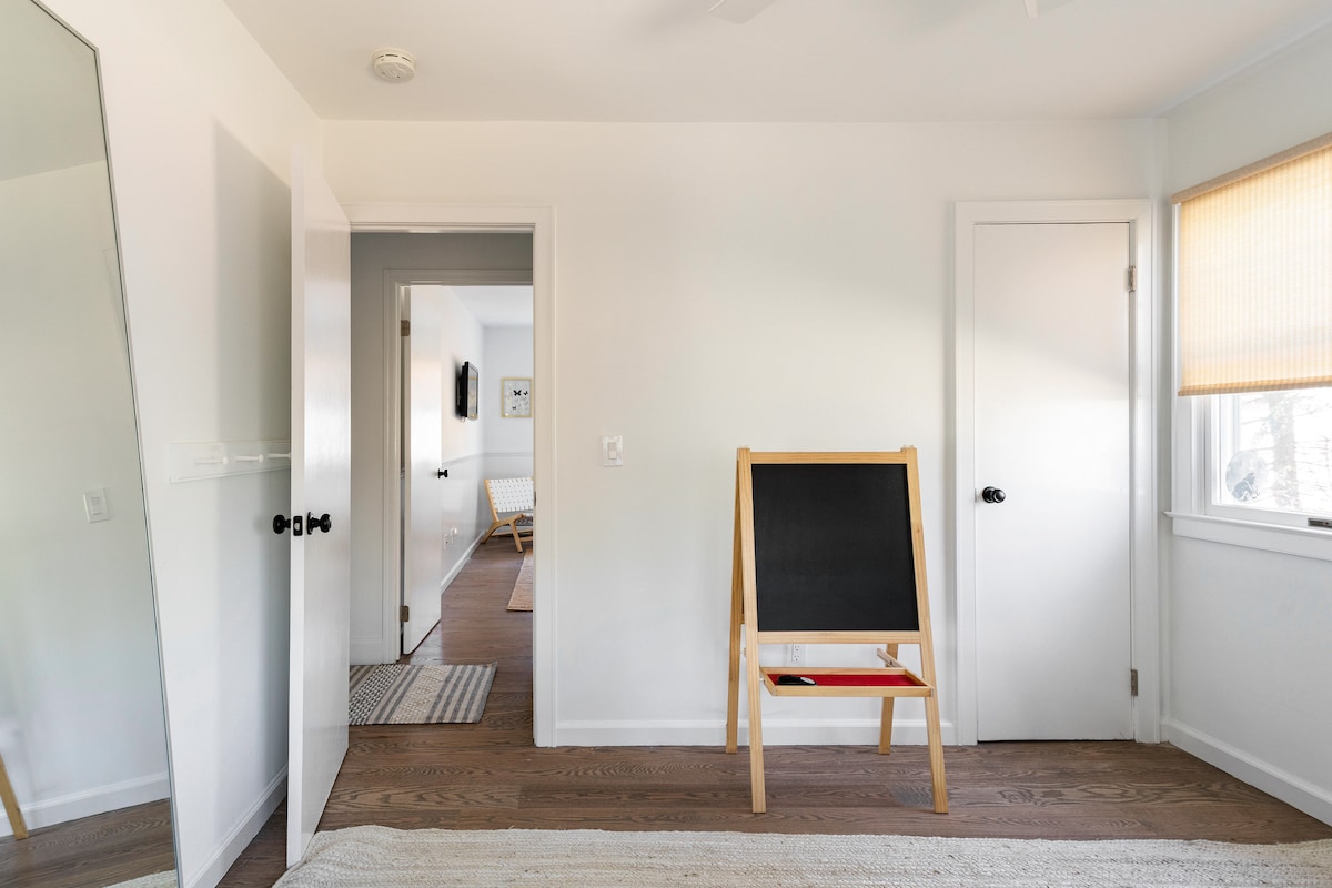 An entryway features a large mirror reflecting light, a small chalkboard easel with a red shelf, and light-colored flooring. A closed door is visible on the right, while an open door leads to another room. Natural light filters through a window covered by a light shade.