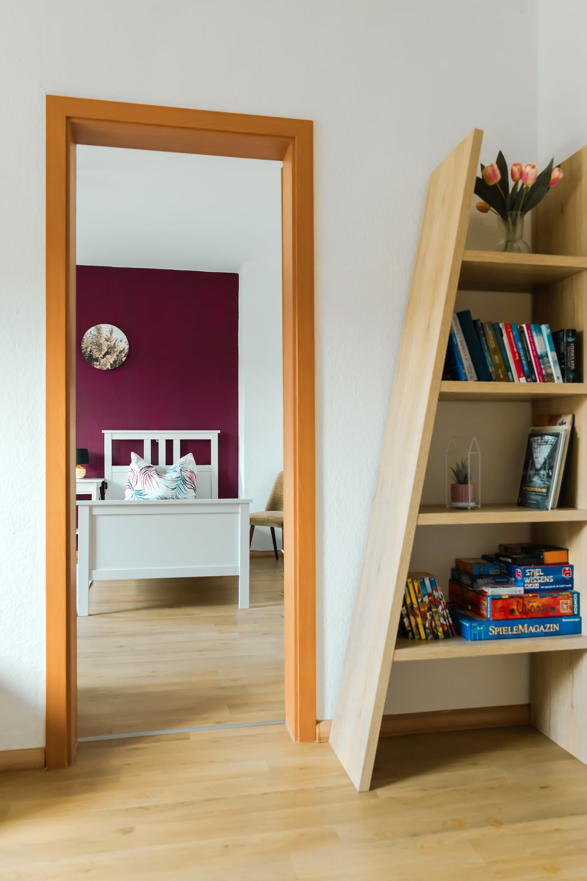 A doorway connects a vibrant room featuring a maroon accent wall and a spacious bed with a decorative pillow. A wooden bookshelf is positioned nearby, displaying books and games. Natural light illuminates the area, enhancing the welcoming atmosphere.