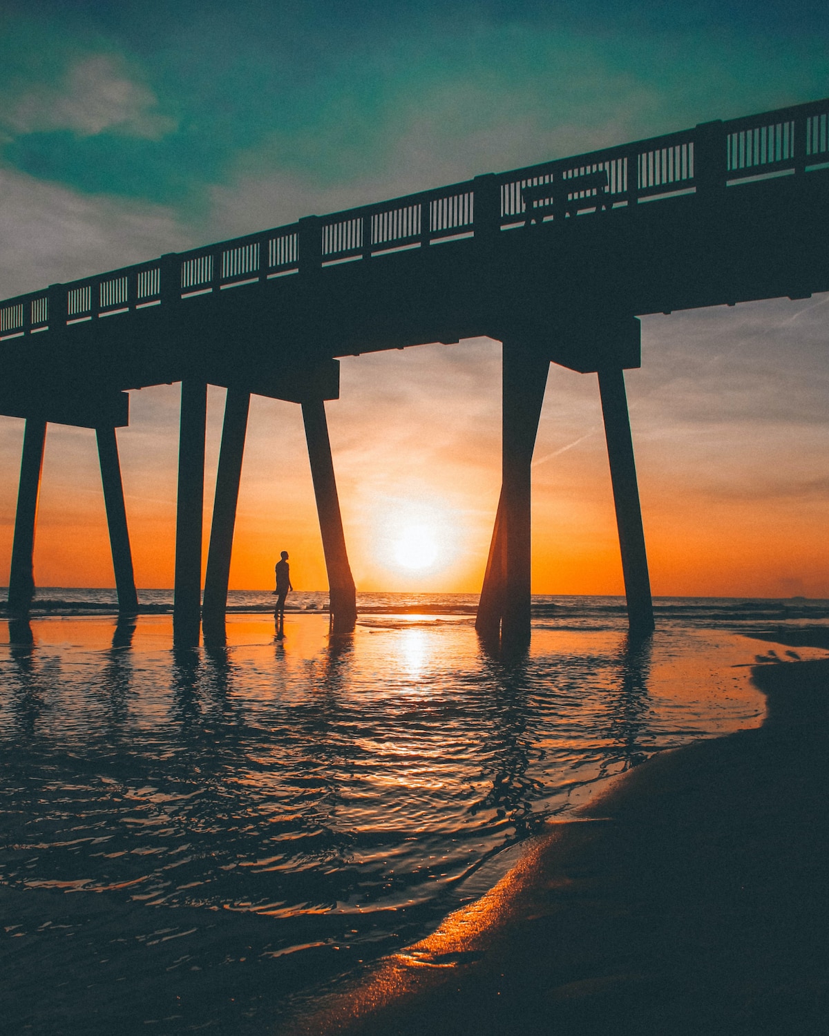 A sunset scene featuring a silhouette of a person walking along a sandy shore beneath a wooden pier. The sky showcases vibrant hues of orange and turquoise, reflecting on the calm water, creating a serene seaside atmosphere.