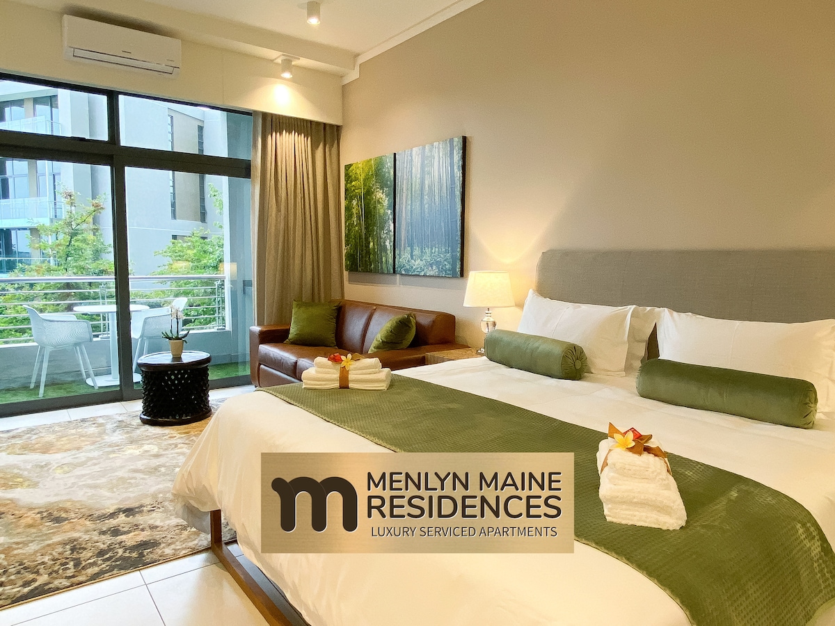 A bright studio is presented, featuring a spacious bed adorned with green accents and neatly arranged towels. A leather couch offers additional seating, while a small table stands nearby. Large glass doors open to a sunny patio, framed by views of landscaped greenery.