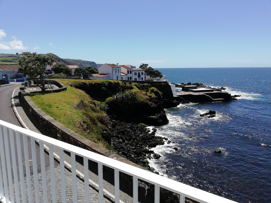 A view of the coastline shows a rocky shore meeting the sea, with gentle waves lapping against the dark rocks. Nearby, a low stone wall runs alongside a paved road, and white buildings are visible on the hilltop, framed by lush greenery and blue sky.