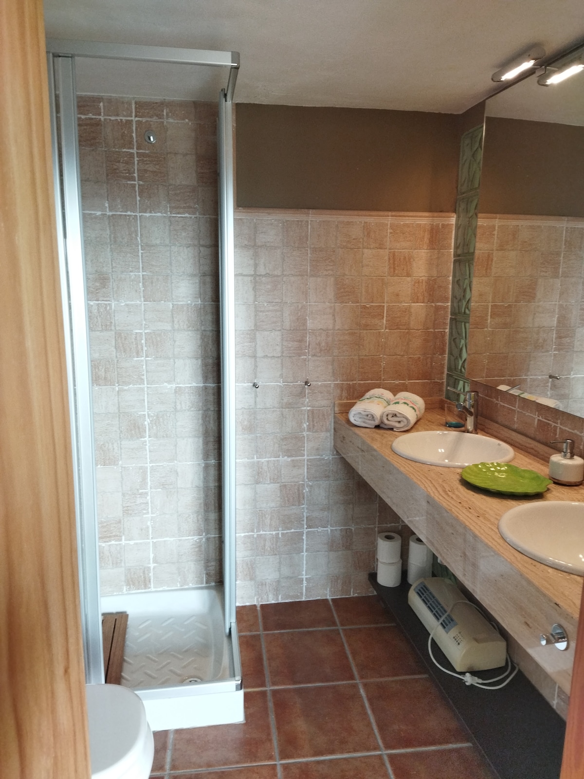 The bathroom features a walk-in shower with glass enclosure and a spacious vanity with two sinks. Neutral tile covers the walls, complemented by wooden accents. Rolled towels are neatly arranged on the counter, and toiletries are positioned for convenience.