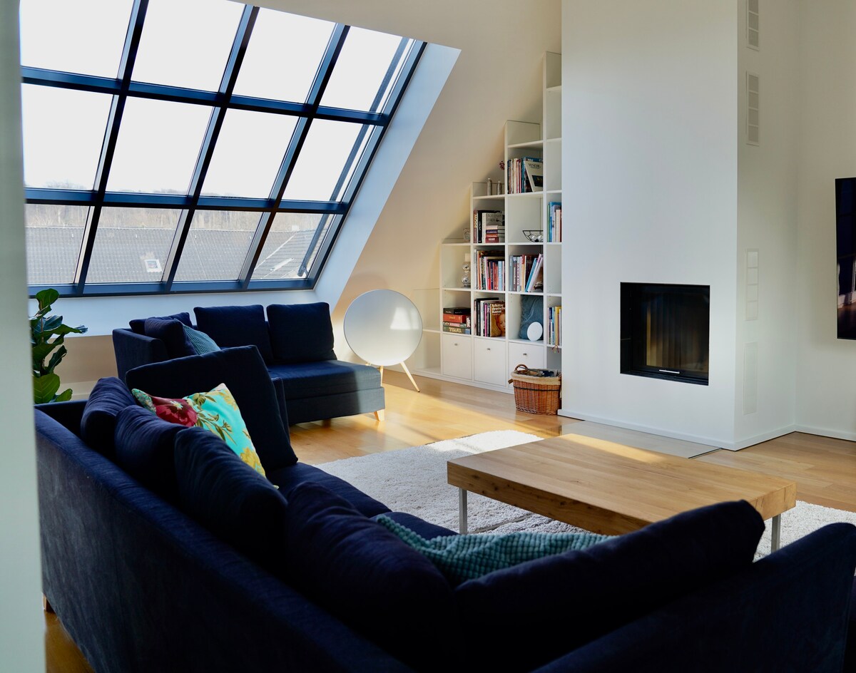 A spacious living area is illuminated by large angled windows that provide natural light. A cozy seating arrangement, including a dark sofa and colorful cushions, is paired with a wooden coffee table. A minimalist bookshelf and a stylish fireplace contribute to the modern decor.