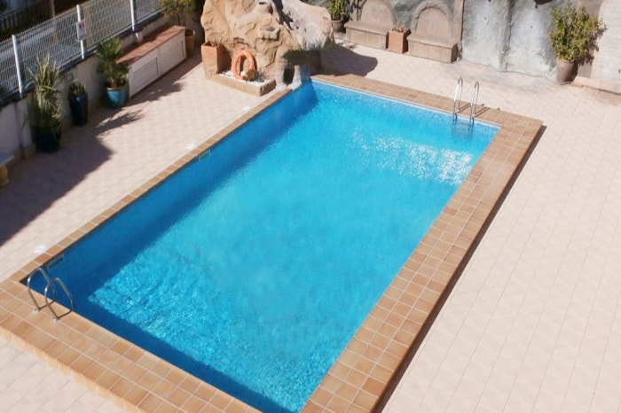 A clear blue swimming pool is framed by a tiled deck, surrounded by potted plants. A decorative rock feature is situated at one end of the pool, enhancing the serene atmosphere. Two ladders lead into the water, available for easy access.
