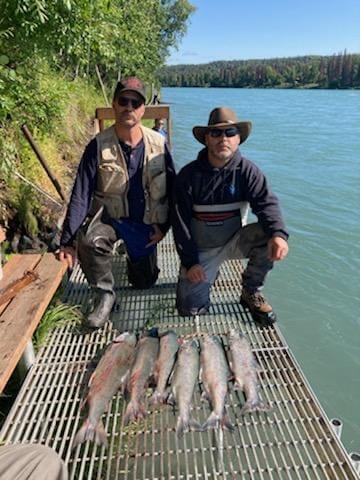 A group is positioned on a metal fishing dock beside the Kenai River, displaying several freshly caught fish arranged neatly on the surface. Lush greenery lines the riverbank, providing a natural backdrop to the fishing experience. The calm water reflects the blue sky and surrounding trees.