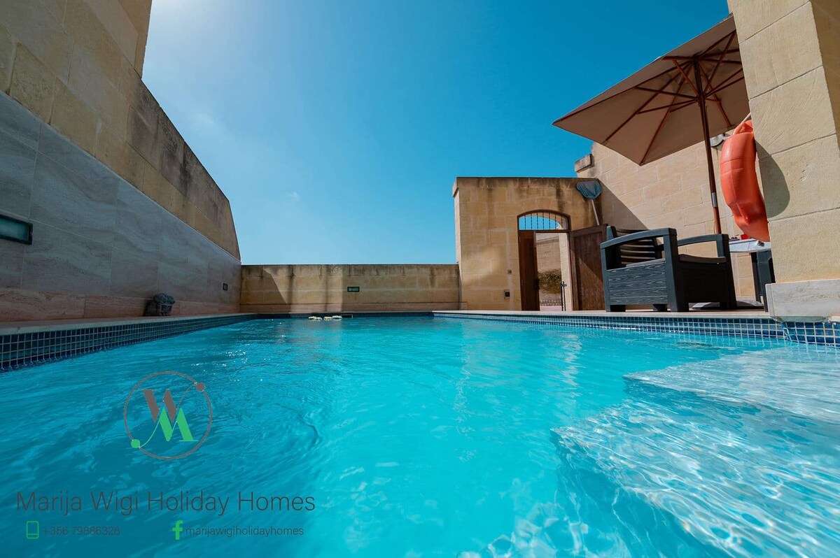 A private pool is surrounded by sleek stone walls, with comfortable outdoor furniture situated nearby. A large parasol provides shade over part of the pool area, while clear blue skies reflect in the water, creating a peaceful and inviting atmosphere for relaxation.
