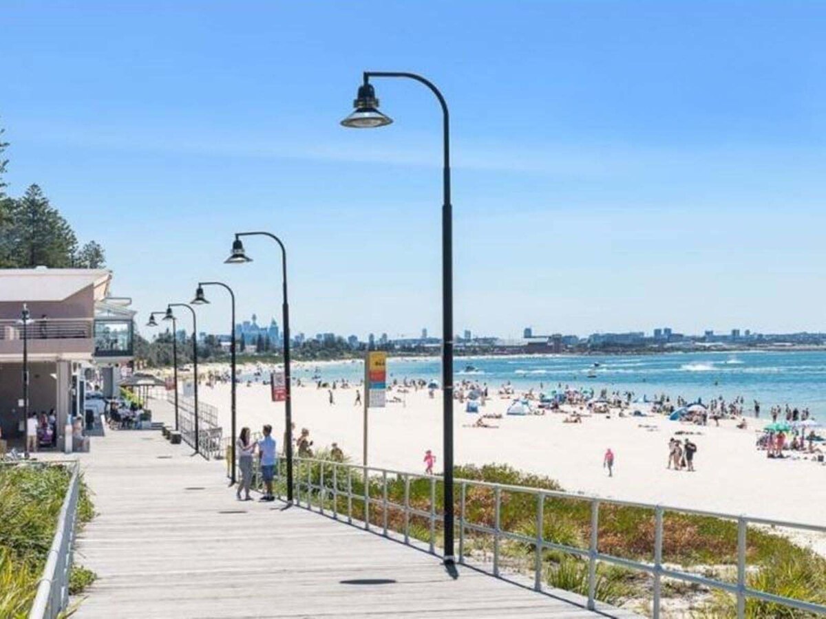 A beachfront promenade is depicted, with a wooden boardwalk lined by stylish lampposts. People can be seen enjoying the sandy beach and water activities under a clear blue sky. Lush greenery borders the walkway, enhancing the coastal atmosphere.