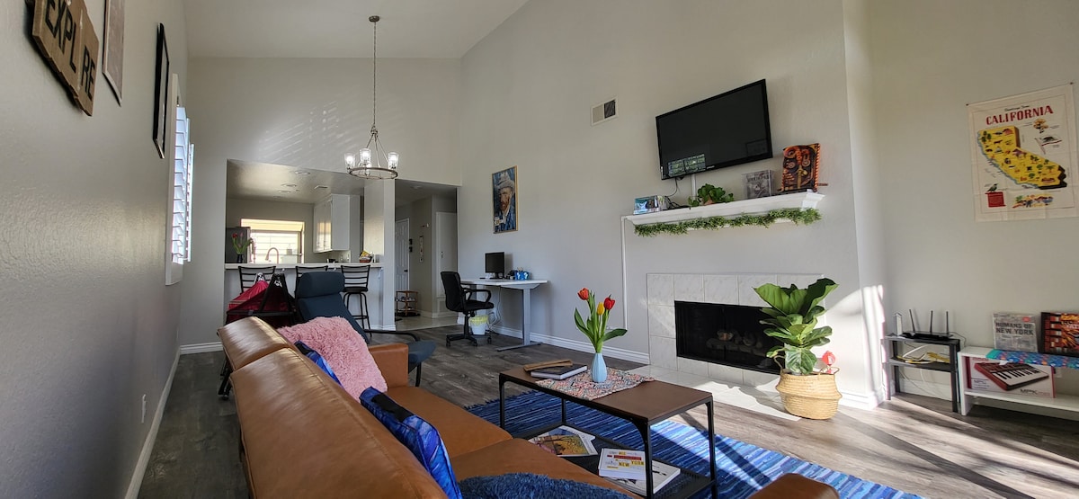 A spacious living room features a comfortable sofa and a modern coffee table, adorned with fresh flowers. A flat-screen TV is mounted above a cozy fireplace, while natural light enters through large windows. The open layout allows a clear view of the adjoining kitchen.