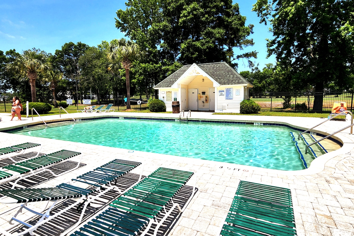A bright outdoor pool area is surrounded by lounge chairs arranged neatly on a tiled deck. The clear water of the pool reflects the sky, and a well-maintained building is visible in the background, providing restroom facilities nearby.