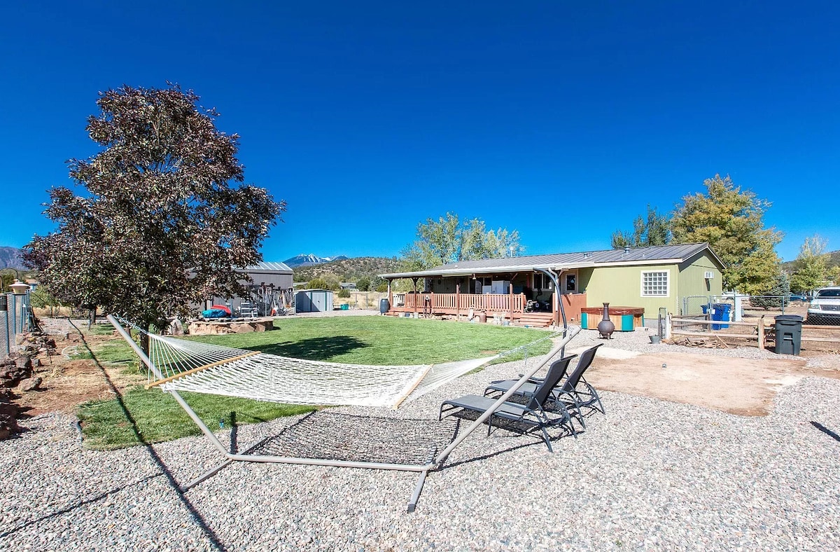 The image displays a spacious backyard, featuring a hammock bed suspended between supports, and two lounge chairs positioned nearby. A well-manicured lawn stretches across the area, bordered by gravel. The home in the background is painted in green, accompanied by trees and a clear blue sky.