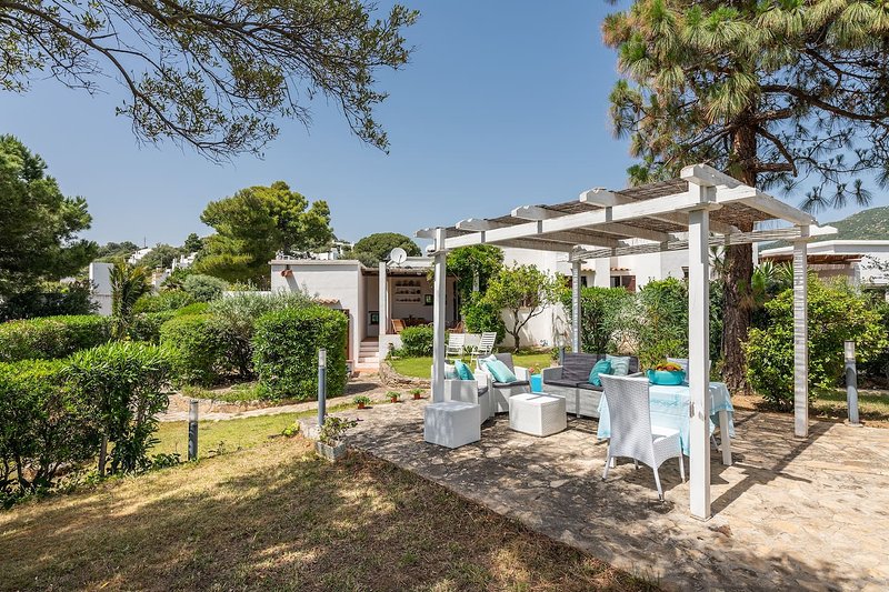 An outdoor patio area is surrounded by greenery, featuring comfortable white seating and a dining table. A pergola offers shade, enhancing the inviting space for relaxation. The well-maintained garden adds to the serene atmosphere of the villa.