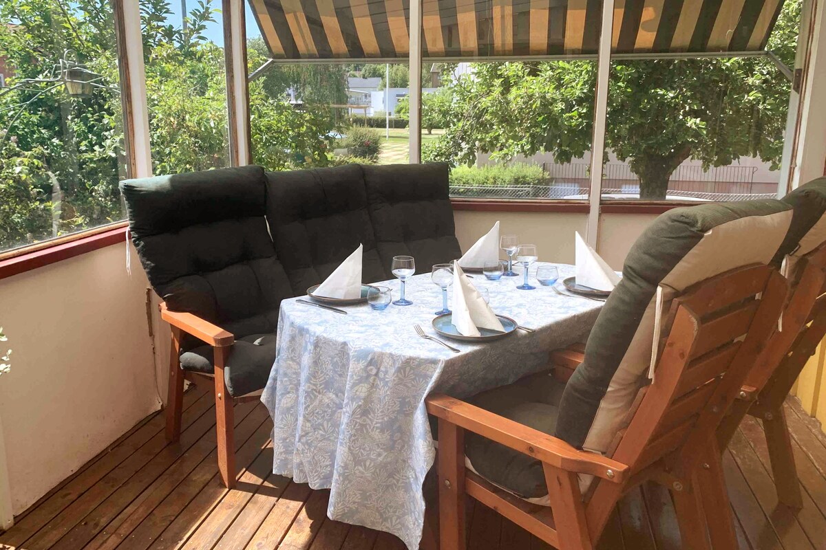 A screened-in dining area is showcased with a wooden table set for four. Plush, upholstered chairs are arranged around the table, while a light blue tablecloth adds a touch of color. Natural light filters through the surrounding windows, highlighting the view of the garden beyond.
