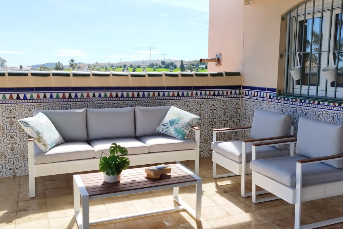 A spacious terrace features modern outdoor seating, including a sofa and two armchairs, accented with cushions. A low wooden table sits in the center, complemented by a small potted plant. Decorative tiled walls provide a vibrant backdrop, and a clear sky is visible through the railing.