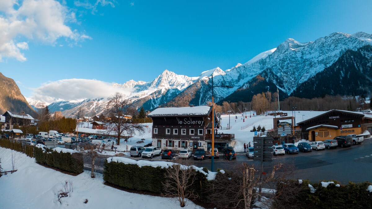 A view of a charming mountain landscape is presented, featuring snow-covered peaks in the background. A quaint building with a wooden façade is visible alongside other structures, with parked cars along a clear pathway. The scene is accompanied by lush greenery and open spaces in the foreground.