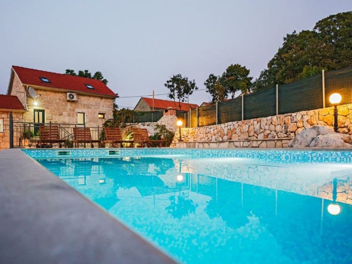 A serene outdoor scene features a large pool, reflecting soft evening lights. Wooden lounge chairs are positioned around the pool, offering a comfortable space for relaxation. The surrounding area showcases natural stone walls and greenery, creating a peaceful atmosphere.