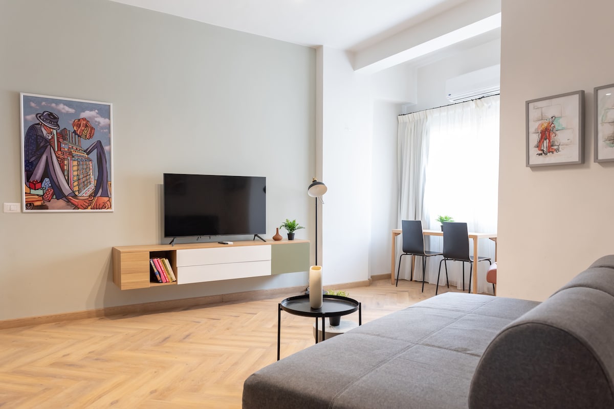 A modern living area features a flat-screen television mounted on a wooden console against a green wall. A gray sofa is positioned to provide comfort. Natural light enters through a large window, illuminating a dining space with two chairs and a table.