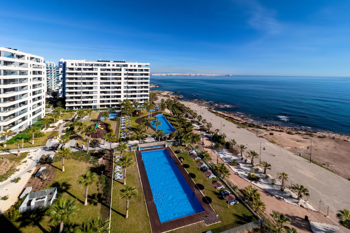 An aerial view captures the expansive grounds of the Panorama Mar complex, featuring two pools surrounded by palm trees. The adjacent beach is visible, with smooth sand meeting the calm sea under a clear blue sky.
