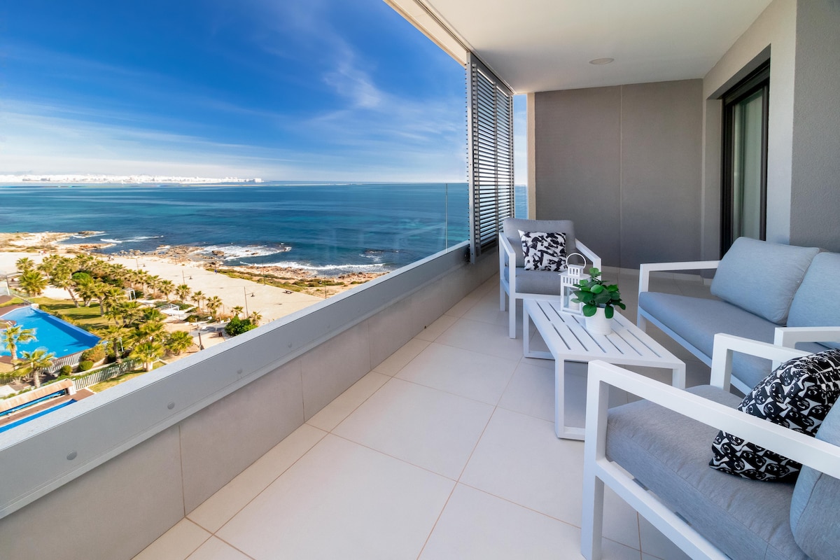 A spacious terrace is presented, featuring comfortable seating arrangements and a small potted plant. The expansive view showcases the coastline, with the sea and sandy beach visible in the distance. A swimming pool is also seen in the landscaped area below.