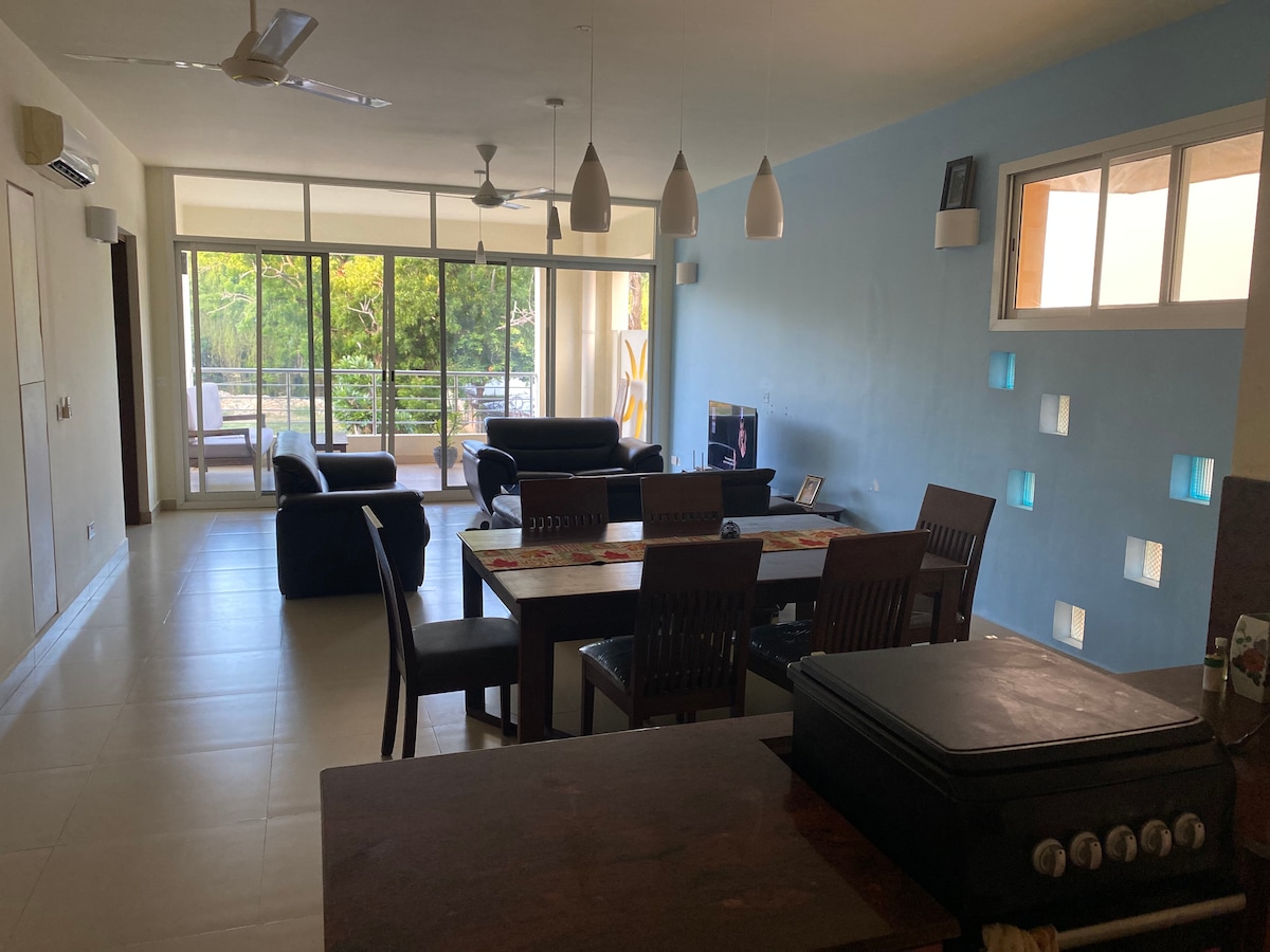 An open living space features a dining table surrounded by chairs, with a comfortable seating area equipped with a television. Large glass doors allow natural light to fill the room, providing views of the balcony and surrounding greenery.