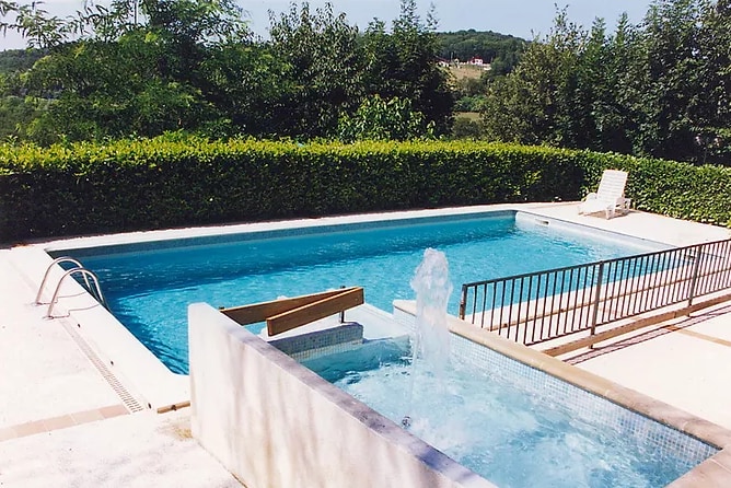 A non-heated swimming pool is surrounded by hedges, with a security fence visible. A seating area is positioned on one side, and a water feature adds a calming element. The tranquil setting emphasizes the property's natural surroundings.