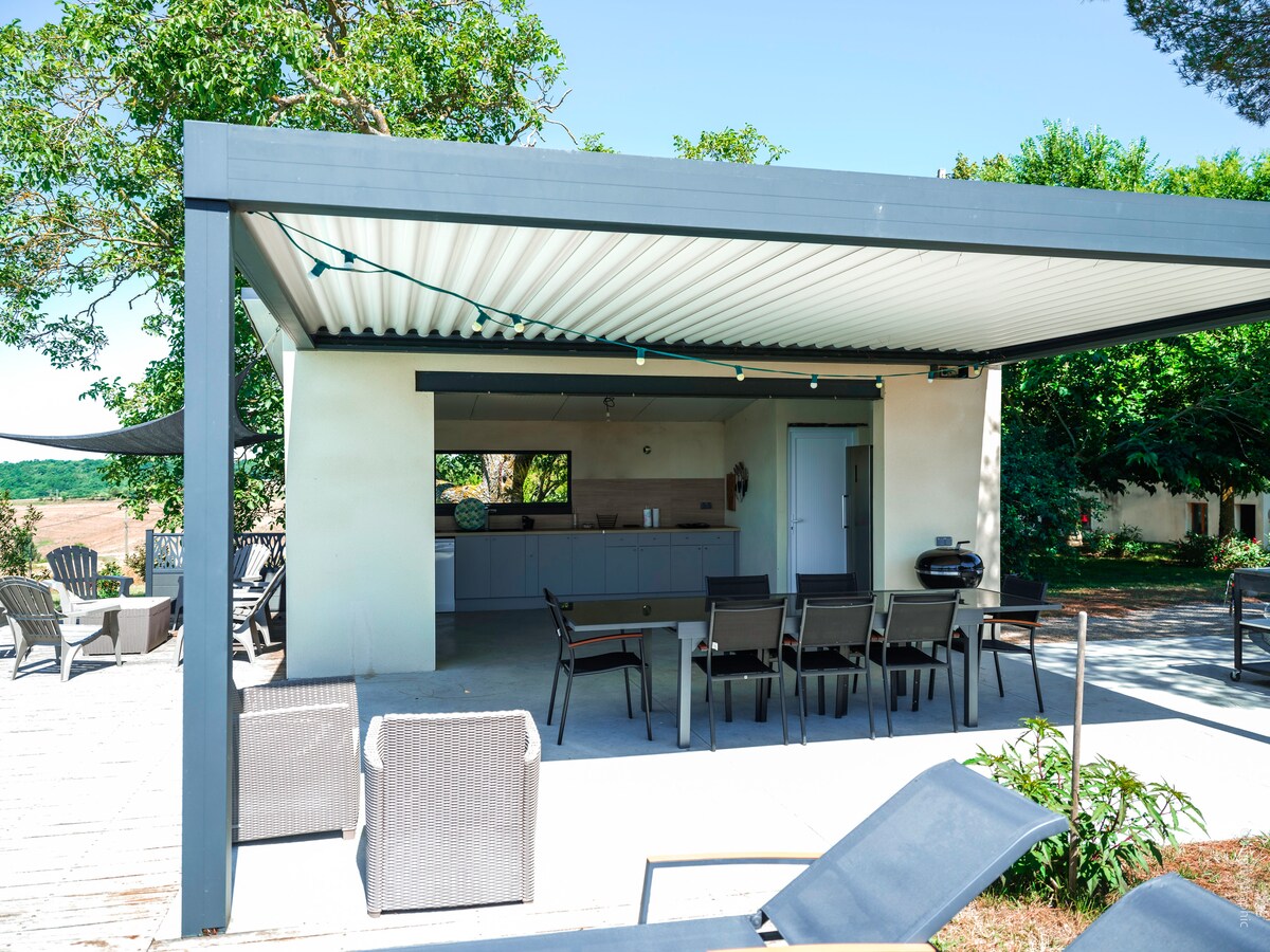 An outdoor pool house features a spacious covered area with a modern kitchen and seating for dining. Relaxation zones are provided with lounge chairs and a barbecue grill, all set against a backdrop of greenery and open farmland.