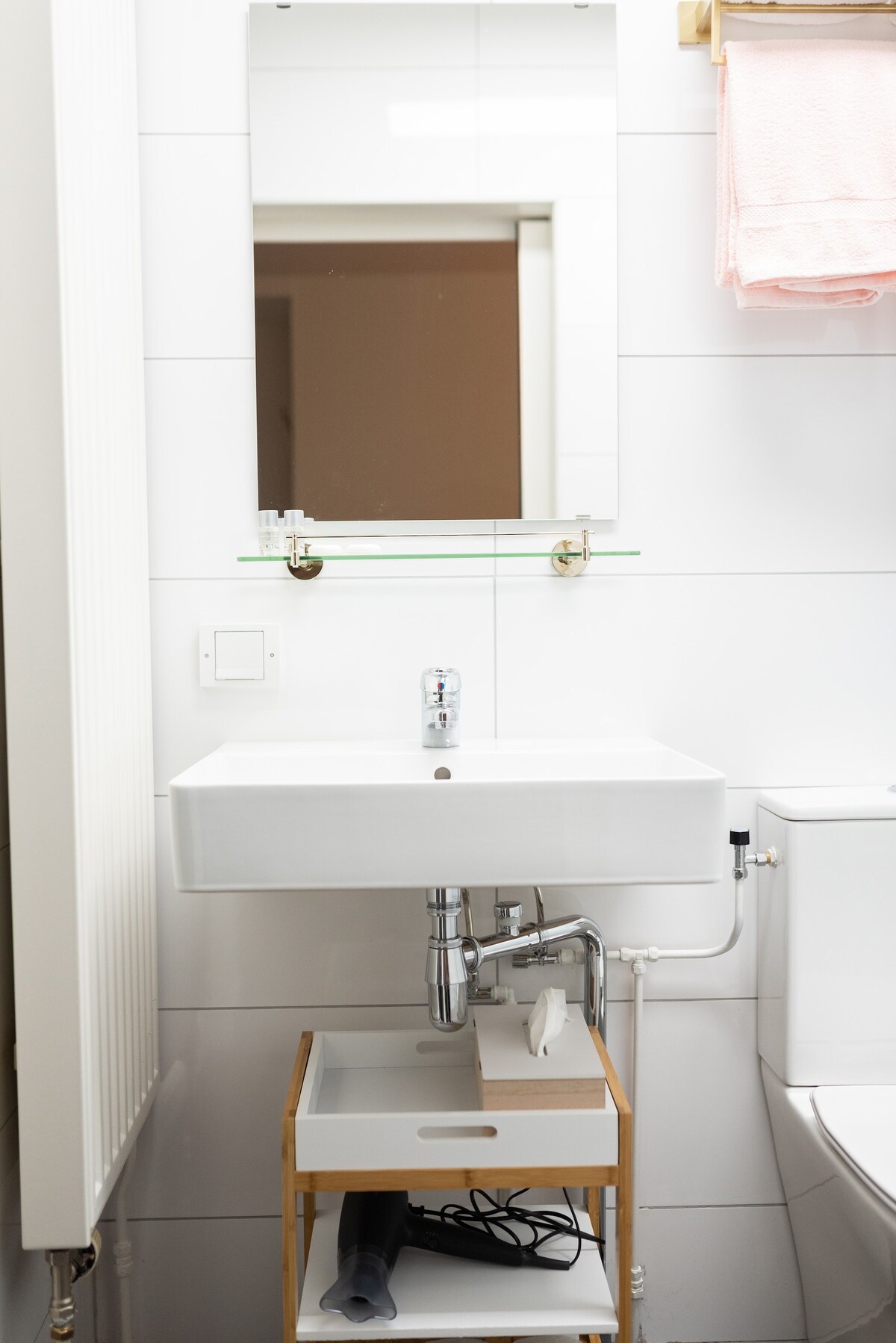 A simple bathroom design is featured, showcasing a modern white sink with a sleek faucet above a bamboo-style cart. A mirror is mounted on the wall above the sink, and a glimpse of a light-colored towel hangs nearby. The white tiled walls provide a clean and bright feel.