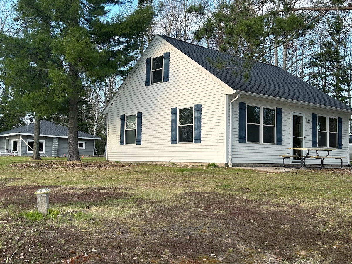 A light-colored cabin is framed by tall trees and a grassy yard. Dark blue shutters accent the windows, and a picnic table is positioned in front, offering an outdoor dining space. A neighboring structure can be seen in the background.