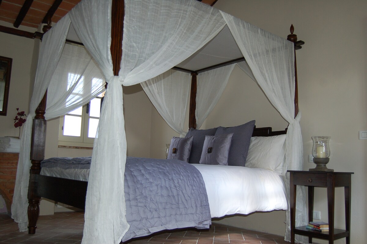 A four-poster queen bed is draped with sheer white fabric, enhancing the serene ambiance of the room. Decorative pillows in muted tones are arranged on the bed. Natural light filters through nearby windows, illuminating the terracotta flooring and original chestnut beams.