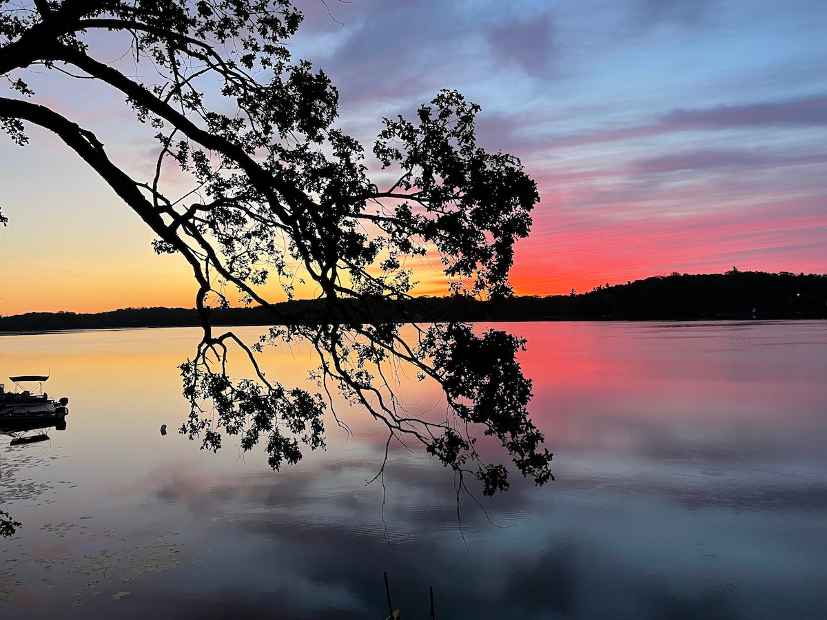 A serene lake view is presented at sunset, with vibrant hues of orange, pink, and purple reflecting on the water's surface. A silhouette of a tree branch extends across the foreground, adding natural framing to the picturesque scene.