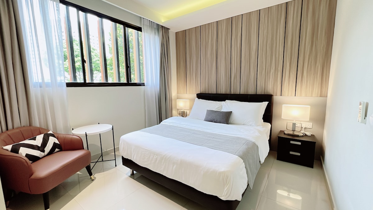 A comfortable bedroom features a queen-sized bed with neutral linens and a decorative pillow. Bedside tables with lamps are positioned on either side, while a cozy armchair and small table are placed near a window with sheer curtains, allowing natural light to brighten the space.