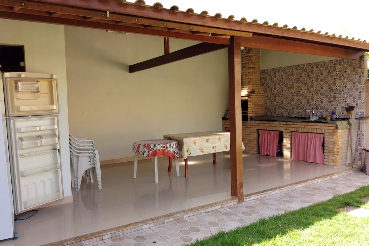 An outdoor barbecue area features a dining table with patterned tablecloth, accompanied by plastic chairs. An outdoor kitchen is equipped with a countertop and grill, while a refrigerator stands nearby. A lawn is visible, enhancing the spacious and open layout.
