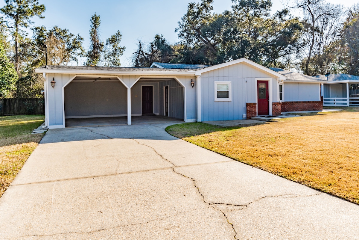 A light blue home features a covered carport and a charming red front door. The driveway leads to a spacious front yard with manicured grass, surrounded by trees in the background. Natural light illuminates the exterior, enhancing the home's welcoming façade.