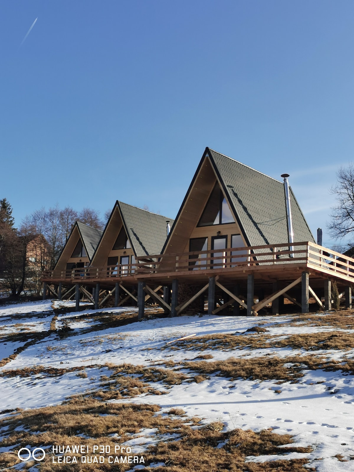 Three A-frame cabins are visible, each set on stilts above a snowy ground. The roofs showcase a dark green hue, complementing the natural surroundings. Large windows are positioned at the front, allowing for ample daylight and views of the landscape.