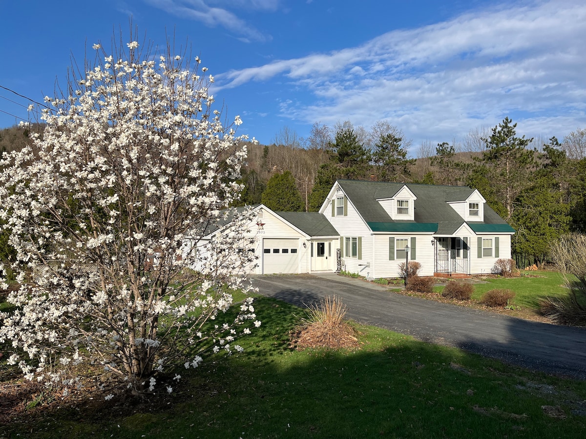 A charming mid-century Cape home is set against a scenic background of rolling hills. A blooming flowering tree frames the front yard, while a gravel driveway leads to an attached garage. The house features a front porch and a well-maintained lawn.