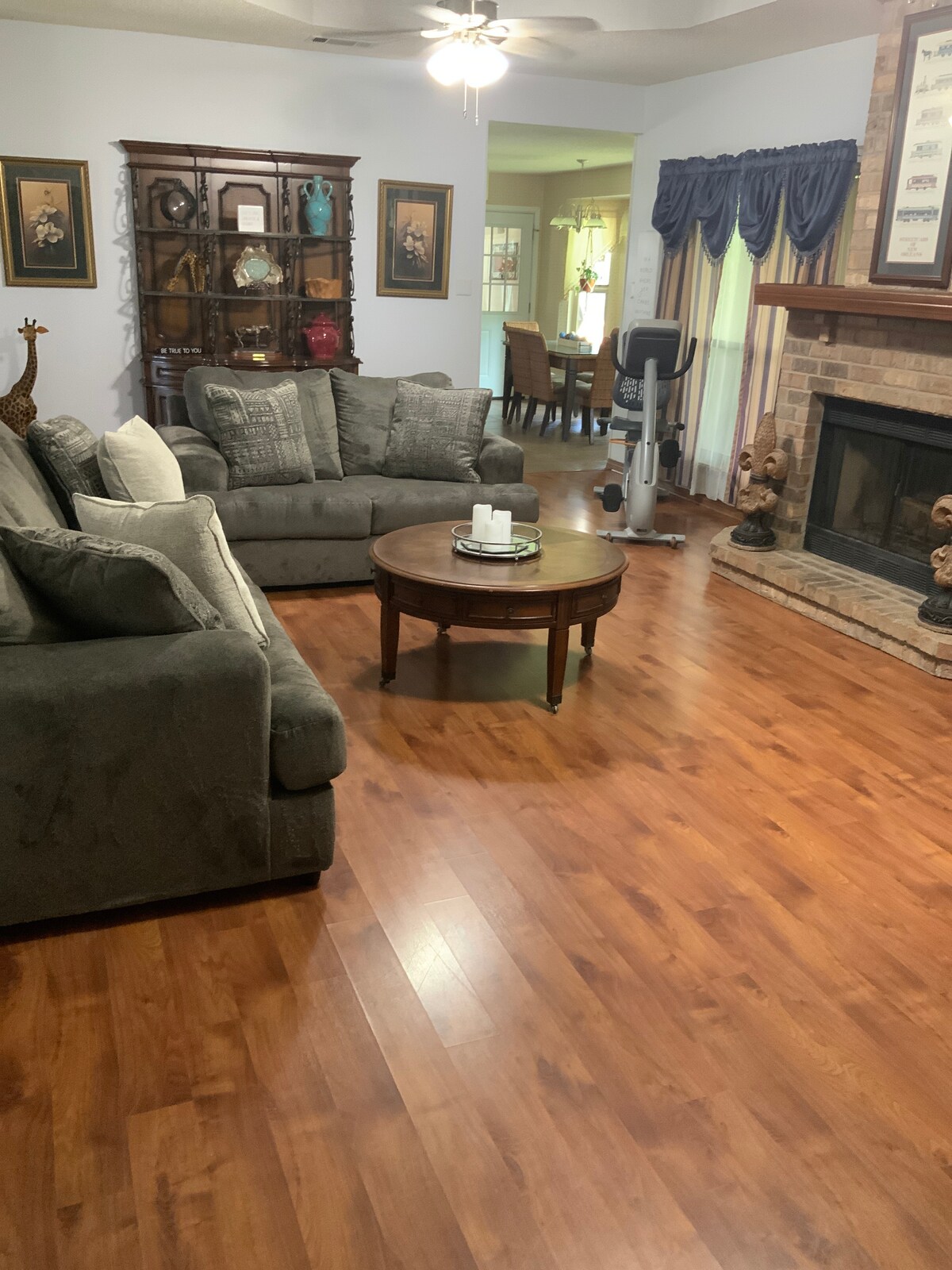 A comfortable living area features plush grey sofas arranged around a circular wooden coffee table. A wooden cabinet displays decorative items, and a stone fireplace adds a cozy focal point. Light wood flooring complements the soft color palette, contributing to the room's inviting ambiance.