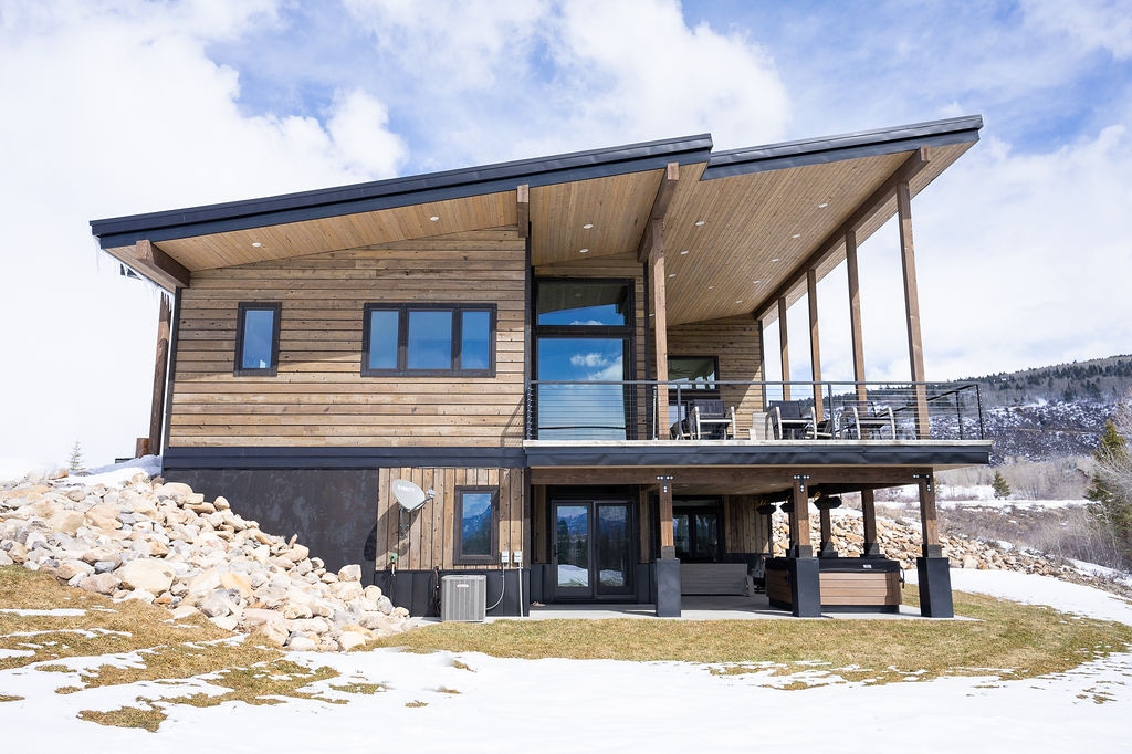 The exterior of a modern mountain retreat is showcased, featuring a combination of wooden paneling and large glass windows. A spacious deck extends from the upper level, overlooking a landscaped area with stone accents and a clear view of the surrounding mountains.